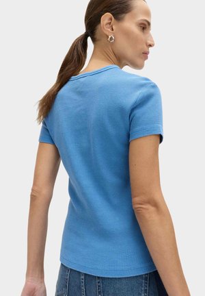 Mujer con cabello castaño recogido en una coleta, que lleva pendientes de aro plateados, una camiseta de manga corta texturizada de color azul claro y vaqueros azules, vista de perfil.