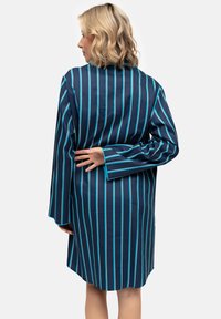 Blondynka zwrócona plecami, ubrana w niebieską, w paski, długosławkową sukienkę, z rękami złożonymi za plecami na jednolitym szarym tle.