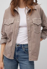 Giacca camicia in corduroy beige con due tasche frontali, chiusura a scatto frontale e colletto a punta, indossata sopra una t-shirt bianca.