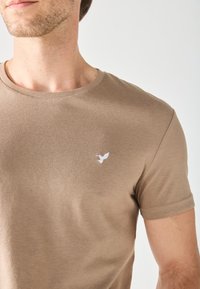 Camiseta de algodón marrón con cuello redondo, mangas cortas y un pequeño logo de un pájaro blanco en el lado izquierdo del pecho. Textura suave y diseño ajustado.