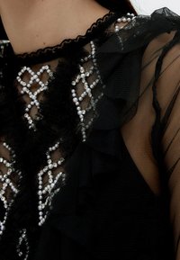 Svart genomskinlig blus med rynkig textur, spetskrage och dekorationer av pärlor och rhinestones i ett geometriskt mönster.