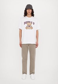 Playera de algodón blanca con un gráfico de un oso y el texto "DOUBLE V" en púrpura; combinada con pantalones beige y zapatillas blancas, rematada con una gorra gris.