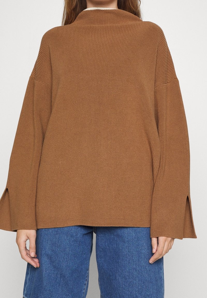 Femme portant un pull marron côtelé ample à manches longues avec des fentes sur les côtés, associé à un jean bleu en denim.