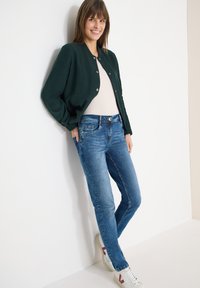 Femme souriante, portant une veste boutonnée vert foncé, un haut blanc, un jean bleu et des baskets blanches, appuyée contre un mur blanc.