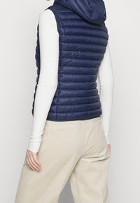 Gilet imbottito senza maniche blu navy con motivo trapuntato orizzontale e cappuccio, abbinato a una camicia bianca a maniche lunghe e pantaloni in velluto a coste color crema.