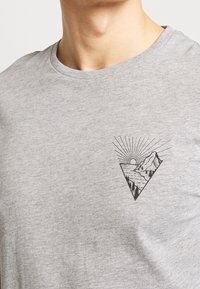 Camiseta de algodón gris con un estampado gráfico negro de montañas, un sol y rayos dentro de una forma triangular en la parte izquierda del pecho.