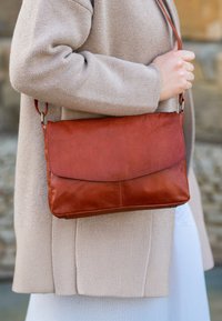 Brun läder crossbody-väska med klaffstängning, rektangulär form, slät yta och justerbar rem. Bärs med en beige kappa.