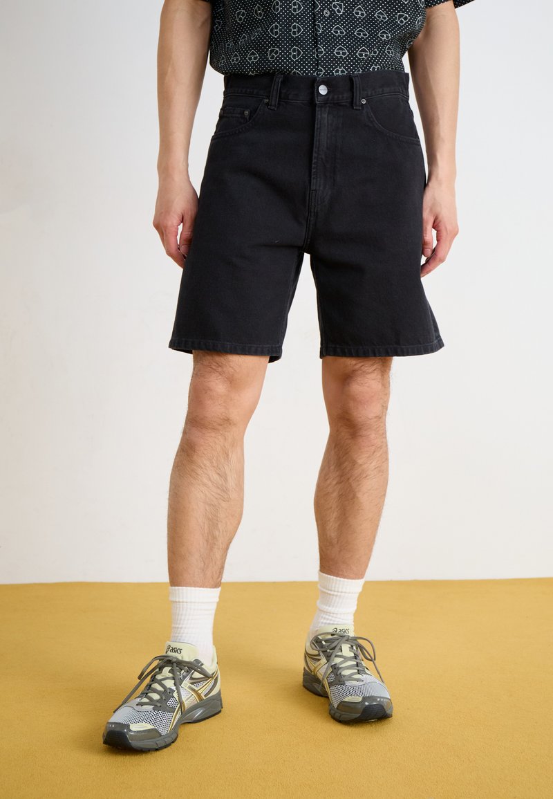 Sort denimshorts med afslappet pasform, fem-lomme-design og syet kant, parret med grå træningssko og hvide sokker.
