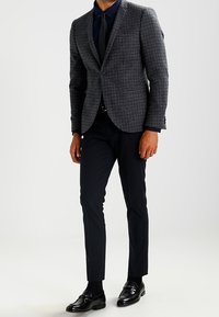 Szary żakiet w kratę houndstooth o dopasowanym kroju, z klapami i pojedynczym zapięciem na guzik, zestawiony z wąskimi granatowymi spodniami i czarnymi butami.
