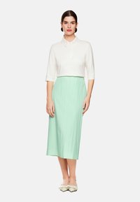 Weißes Blusenshirt mit besticktem Kragen und Dreiviertelärmel, kombiniert mit einem plissierten, mintgrünen Midirock und spitzen, weißen Ballerinas.