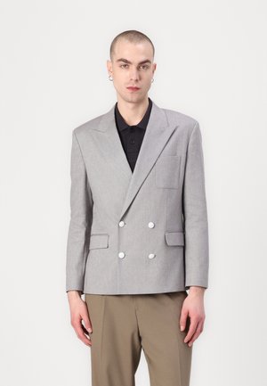 Grau doppelt knöpfiges Blazer mit Revers, weißen Knopfdetails und einer Fronttasche. Getragen über einem schwarzen Poloshirt.