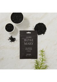 Emballage noir pour "Pack Équilibrant de Scalp au Cold Brew et Romarin" avec charbon. Entouré d'échantillons de poudre noire et de feuilles vertes sur une surface claire.