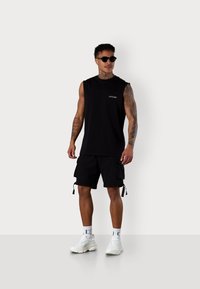 Schwarzes ärmelloses Baumwollshirt mit Logo, kombiniert mit schwarzen Cargo-Shorts mit Seitentaschen und weißen klobigen Sneakers.