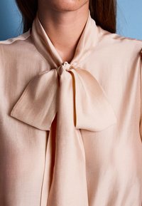 Gaisma persiku krāsas satīna blūze ar sasietu loku kaklā, ar gludu tekstūru un minimālistisku dizainu.