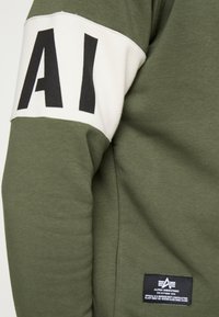 Felpa verde oliva con polsini bianchi e lettere nere "AI". Include un'etichetta nell'angolo in basso a destra con i dettagli del marchio.
