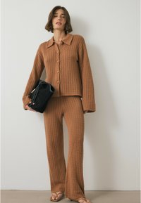 Bruin gebreid set bestaande uit een colbert met knopen en een col en wijde broek met open geweven textuur. Model draagt een zwarte handtas.