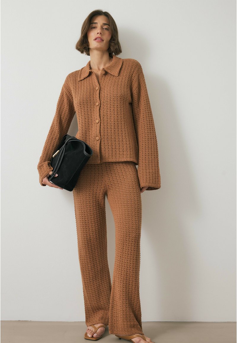 Bruin gebreid set bestaande uit een colbert met knopen en een col en wijde broek met open geweven textuur. Model draagt een zwarte handtas.