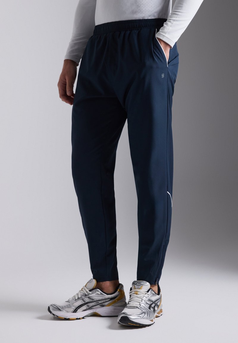 Navy athletic pants with elastic waistband and pockets, featuring a tapered shape and minimal branding. Worn with silver running shoes.