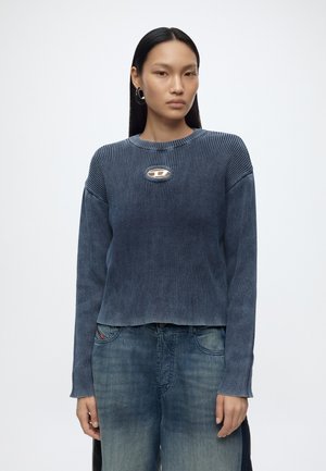 Femme aux longs cheveux noirs portant un pull côtelé bleu avec une petite découpe ovale au niveau de la poitrine et un jean bleu délavé, debout sur un fond blanc.