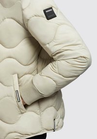 Vadderad jacka i ljus beige med vågig kviltad textur, ribbade muddar och en svart logotyppatch på axeln. Sidoficka synlig.