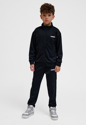 Pojke som står och bär en svart Hummel-träningsdräkt med en zip-up jacka och matchande byxor, samt grå höga sneakers.