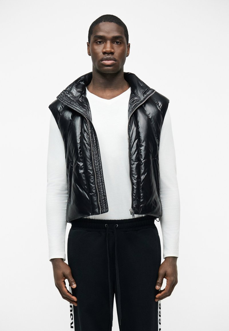 Άνδρας που φοράει μαύρο, γυαλιστερό γιλέκο χωρίς μανίκια πάνω από λευκό πουκάμισο με μακριά μανίκια και μαύρο παντελόνι με κορδόνια στη μέση και κείμενο στο πλάι.