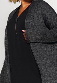 Cardigan grigio a maglia con apertura frontale, tessuto leggero e maniche lunghe, indossato sopra una maglietta nera con chiusura a zip sul collo.