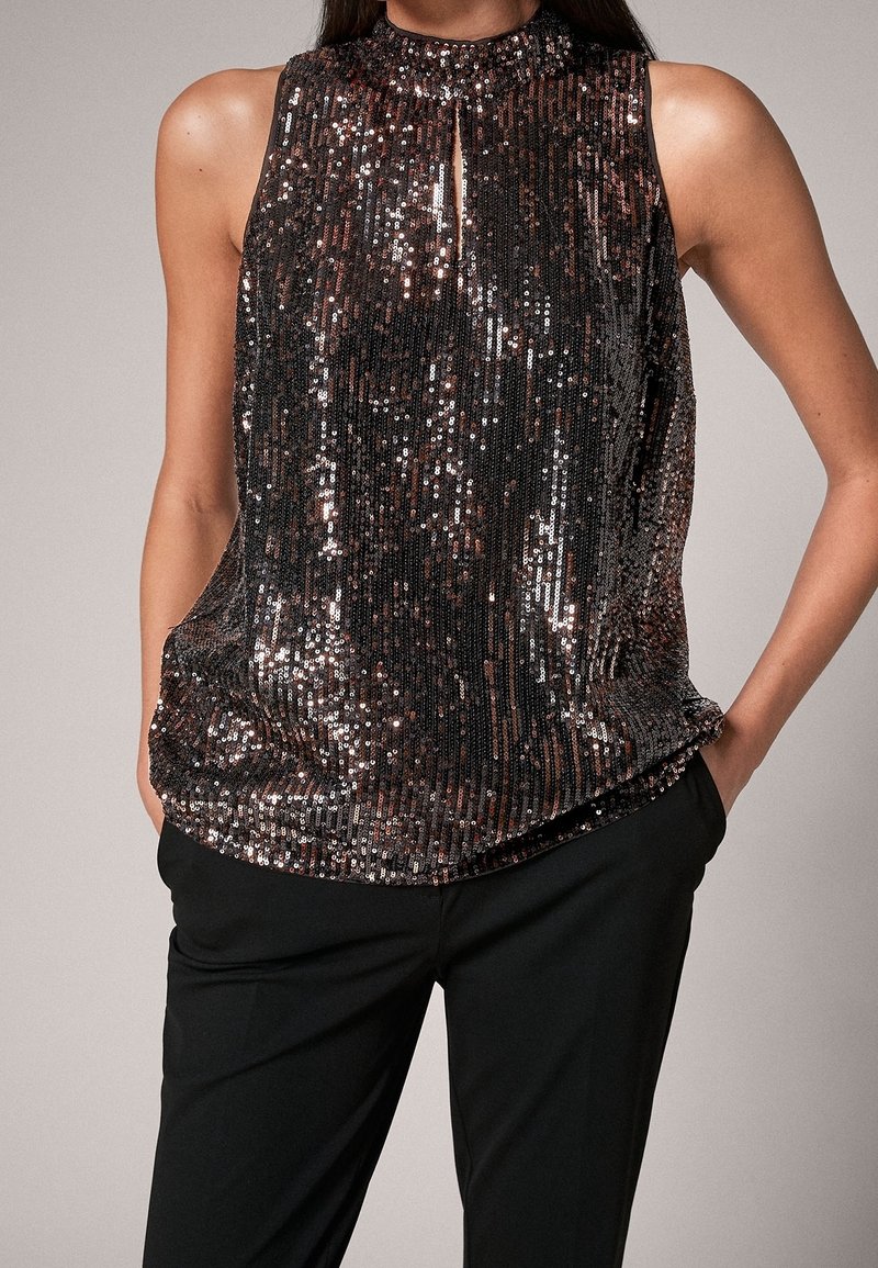 Femme portant un top sombre sans manches à sequins avec une petite fente à l'avant, associé à un pantalon noir, mains dans les poches, sur fond uni.