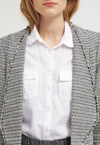 Blazer en diseño de espiga en tonos gris y blanco con bordes deshilachados, usado sobre una camisa blanca con botones que tiene dos bolsillos en el pecho y una falda gris.