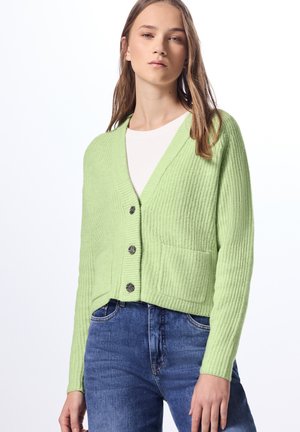 Cardigan verde chiaro con texture a coste, scollo a V e bottoni frontali. Presenta due tasche frontali e lunghezza cropped. Indossato sopra una camicia bianca.