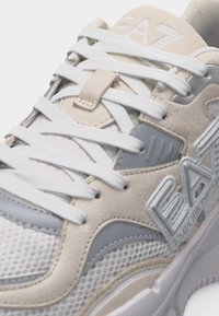 Μπεζ και λευκό αθλητικό παπούτσι με λευκά κορδόνια, υφές από δίχτυ και σουέντ, με ένα εμφανές λογότυπο BA7 Emporio Armani στο πλάι.