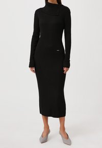 Robe noire en tricot côtelé avec un col haut et des détails de boutons. Manches longues et silhouette ajustée, atteignant le genou. Texture lisse.