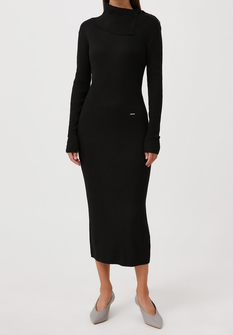 Robe noire en tricot côtelé avec un col haut et des détails de boutons. Manches longues et silhouette ajustée, atteignant le genou. Texture lisse.