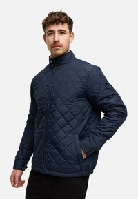 Marineblauer gesteppter Mantel mit hochgeschlossenem Kragen, Reißverschluss und seitlichen Taschen. Verfügt über ein Rautenmuster und Druckknopfakzente am Kragen.