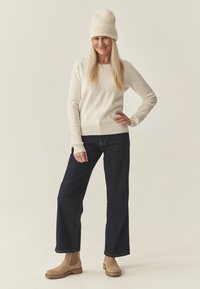 Pulover crem cu un design embosat de urs, blugi închiși la culoare și cizme până la gleznă de ton deschis. Textură cu nervuri pe manșetele puloverului și o pălărie tricotată asortată de culoare crem.