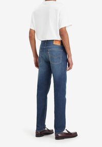 Jeans en denim bleu avec une coupe droite, dotés d'une patch en cuir à l'arrière et d'un design classique à cinq poches. Portés avec un t-shirt blanc.