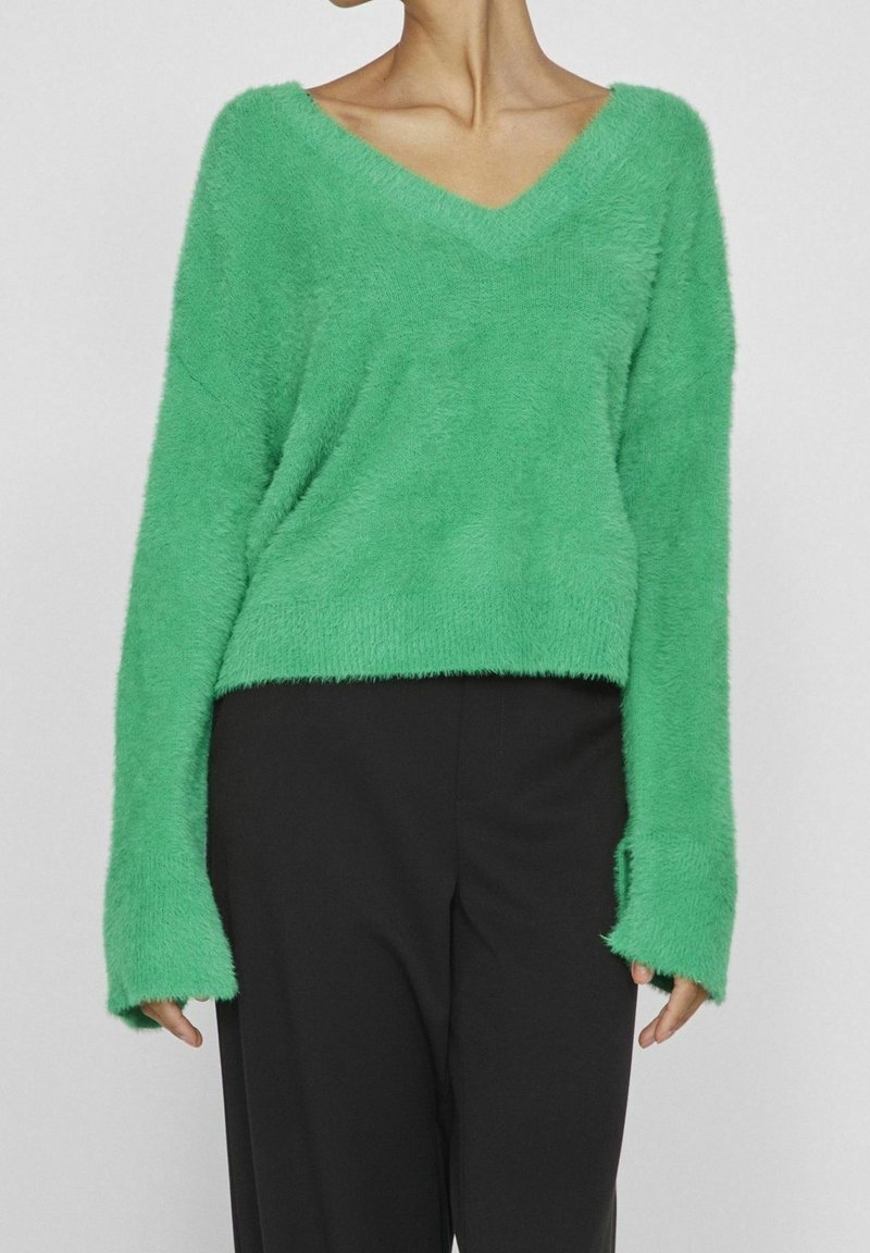 Femme portant un pull vert vif en maille duveteuse à col en V avec des manches longues et un pantalon noir, montrée du cou aux hanches sur un fond uni.