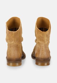 Par de botines tobilleros de ante color beige vistos desde atrás con detalles de costura y suelas de goma marrón sobre un fondo blanco.