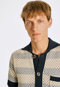 Stickad polo i randigt nätmönster med beige, grå och marinblå färger. Har en ribbad krage och knäppning med en bröstficka.