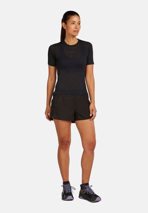 Kvinde stående, iført sort kortærmet tætsiddende skjorte, sorte shorts og sorte vandresko med lilla snørebånd, på en ensartet lys baggrund.