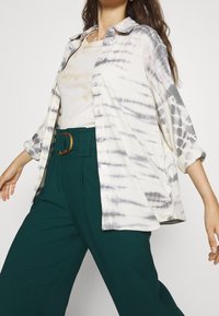 Knappad skjorta i kräm- och grått tie-dye-mönster, ihop med högmidjade gröna byxor med en rund bältesknäpp av trä.