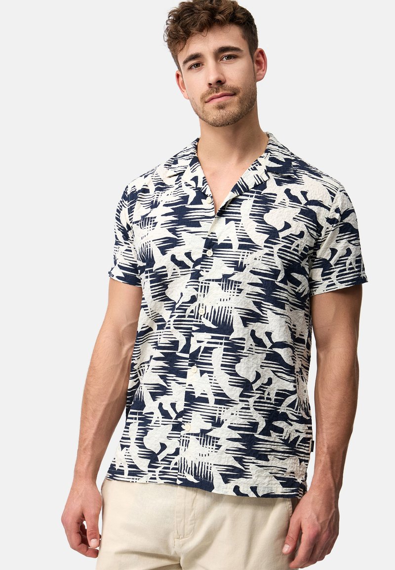 Camisa de manga corta en tejido ligero con un patrón abstracto en azul y blanco; presenta un cuello de tipo camp collar y un cierre frontal con botones.