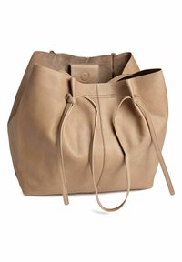 Beige bőr tote táska puha textúrával, összekészített tetejével, két hosszú pánttal és kör alakú fém díszítéssel a zárón.