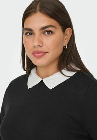 Crna pulover s bijelim, točkastim ovratnikom. Glatka tekstura, kroj koji se prilagođava tijelu i okrugli izrez. Naušnica vidljiva na modelu.