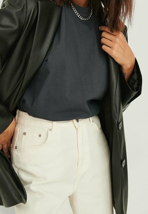 Blazer di pelle nera sopra una T-shirt grigio scuro, abbinato a jeans bianchi a vita alta. Una collana a catena argentata aggiunge dettaglio. Texture lisce.