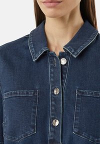 Denimhemd in einem dunklen Blauton, mit Knopfverschluss und metallischen Akzenten sowie zwei Brusttaschen. Klassisches Kragen-Design und glatte Textur.