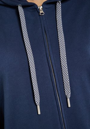 Le hoodie zippé bleu marine présente un tissu texturé, une fermeture éclair argentée, ainsi que des cordons de serrage contrastants blancs et bleu marine avec des embouts en métal.
