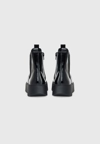 Bottines en cuir verni noir avec une semelle épaisse, fermeture éclair et tirette au talon. Texture lisse et design épuré.