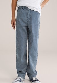 Isik, kes kannab sinise ja valge vertikaaltriibuliste teksapükstega, valget T-särki ja tumelsiniseid tossusid valgete paeltega, lihtsa tausta ees.