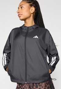 Sort letvægts lynlåstrøje med hætte, med hvidt Adidas-logo og tre stribede detaljer på ærmerne. Glat, skinnende tekstur.